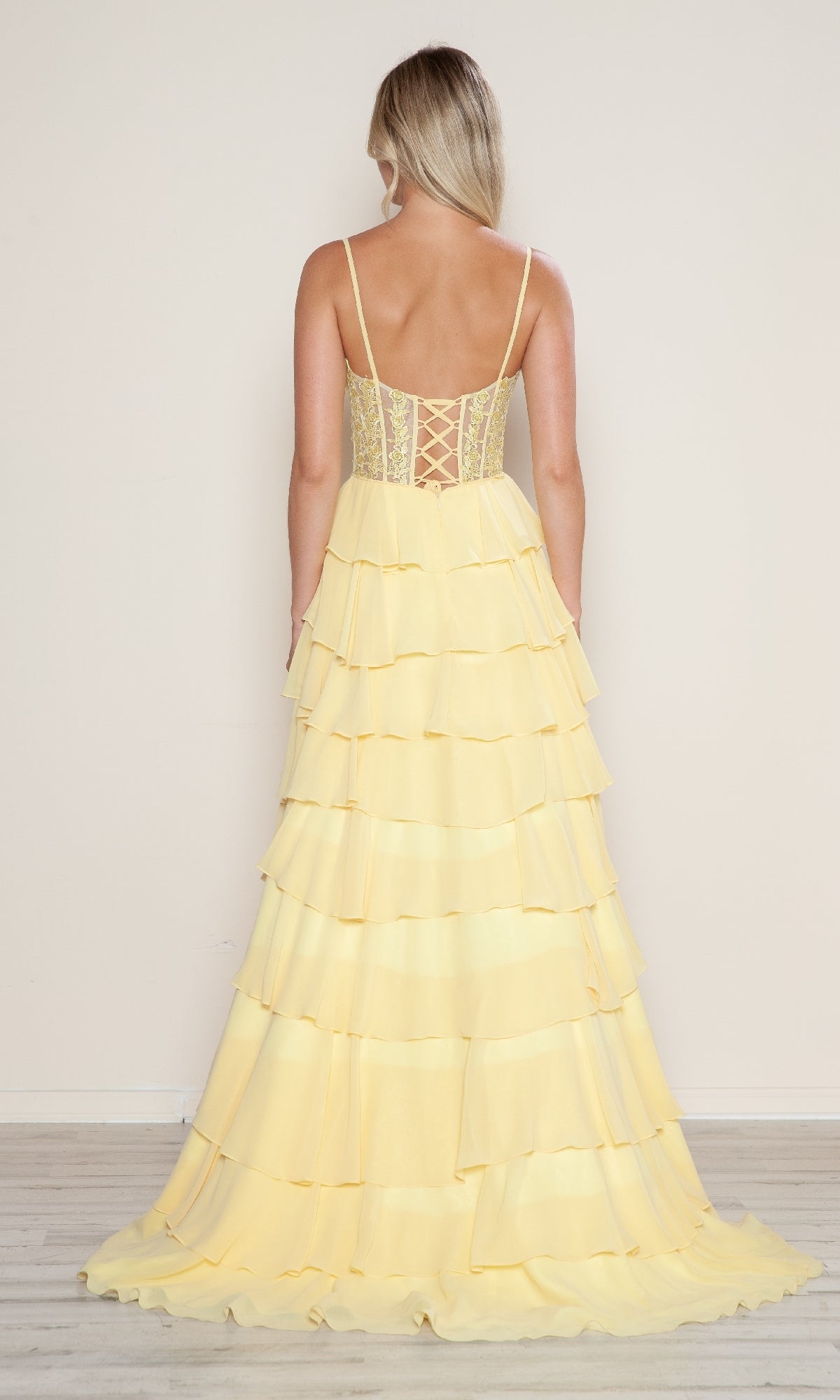 Woman with her back to the camera wears the Poly USA 9698 Long Tiered Corset Prom Dress, featuring a light yellow A-line silhouette, spaghetti straps, and lace-up corset-style bodice detail at the back.